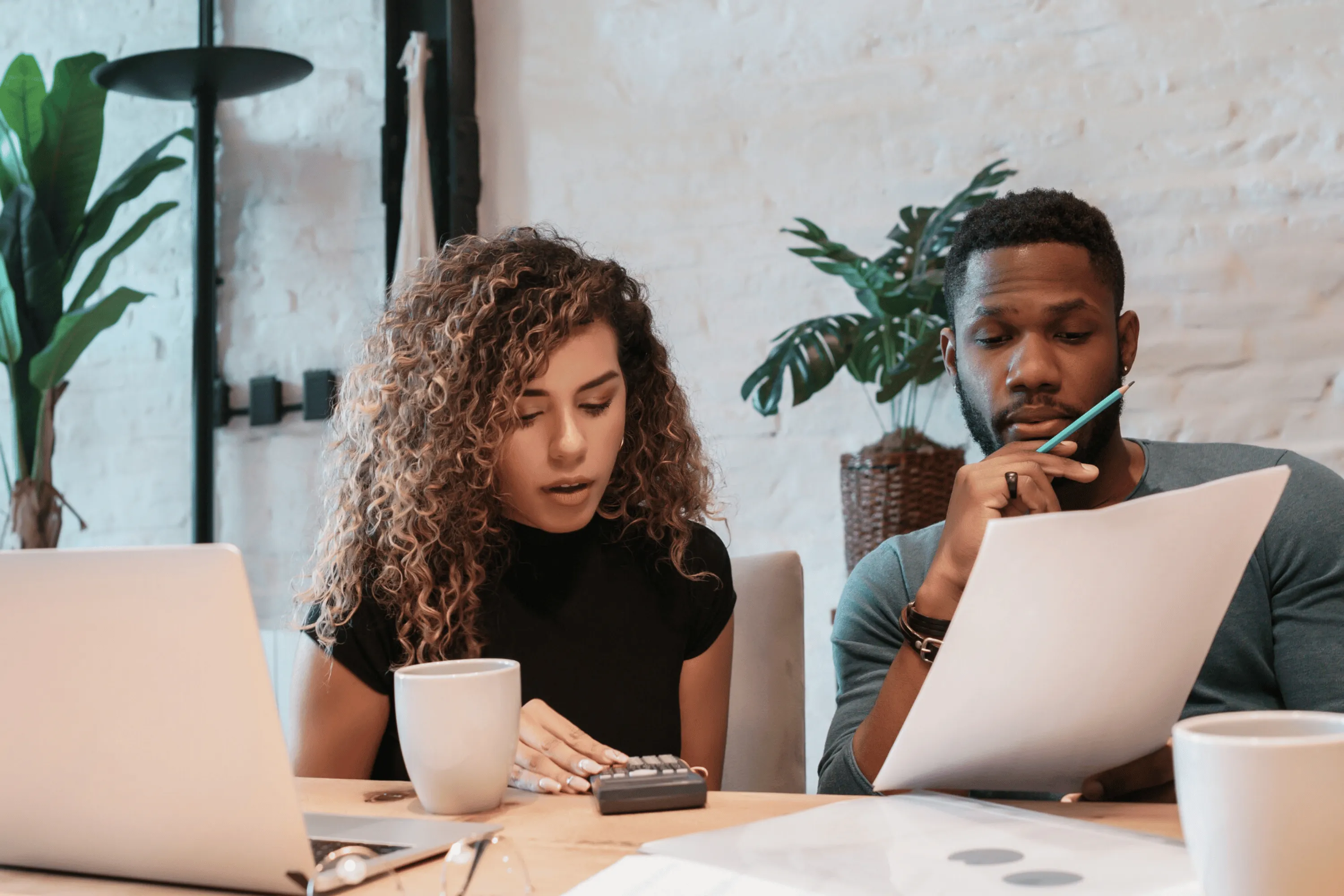 A couple planning together at a table, laptops, papers and coffee in front of them.
